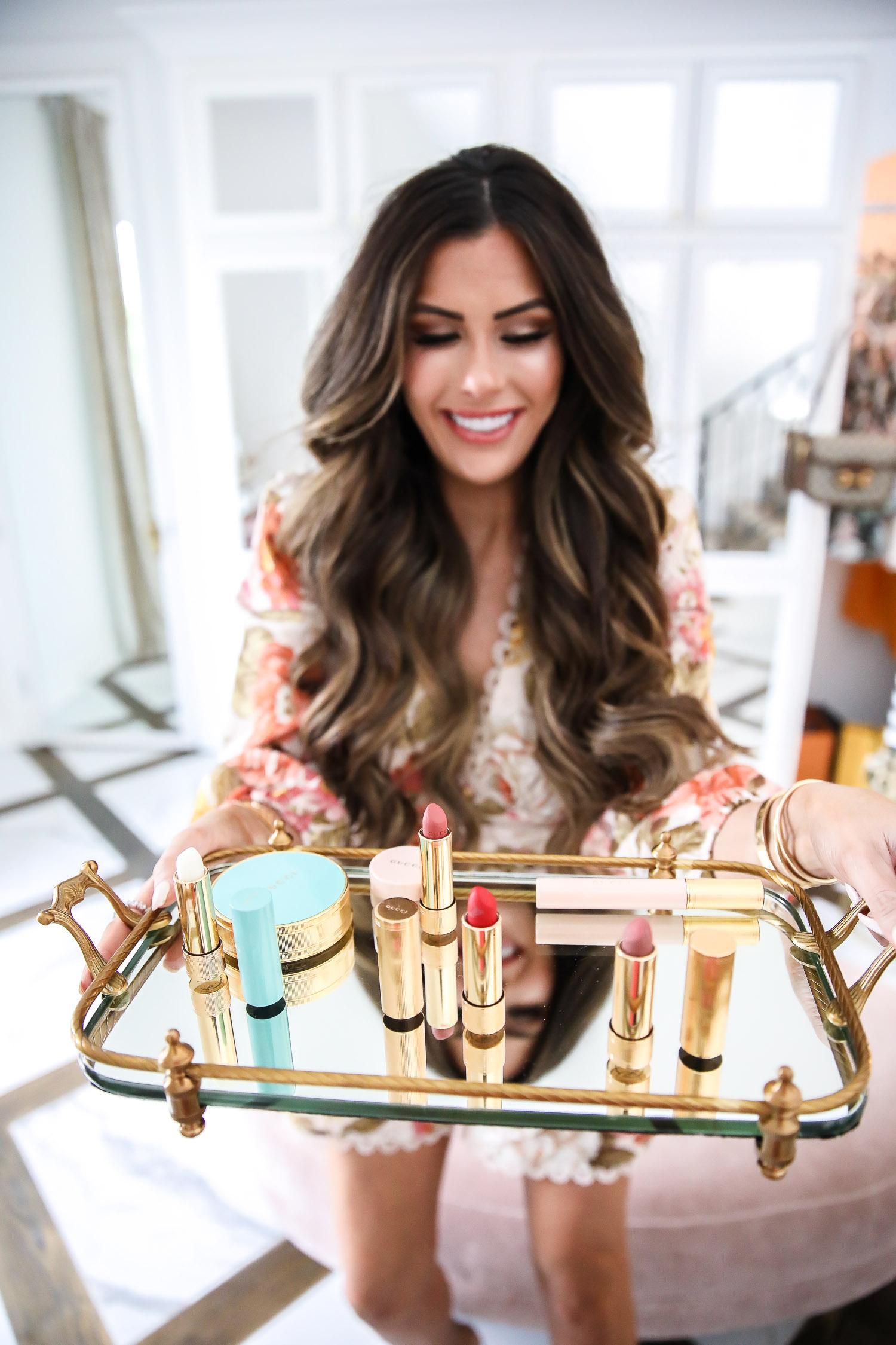 gucci bronzer review, new gucci makeup 2020, gucci lipstick 2020, gucci lip balm, top beauty blogger 2020, zimmerman romper, emily gemma-22 | Gucci bronzer review by popular US beauty blog, The Sweetest Thing: image of Emily Gemma wearing a floral romper and sitting on a tuft velvet ottoman while holding a tray containing Gucci Poudre De Beauté Éclat Soleil Bronzing Powder, Gucci mascara, and Gucci lipstick.