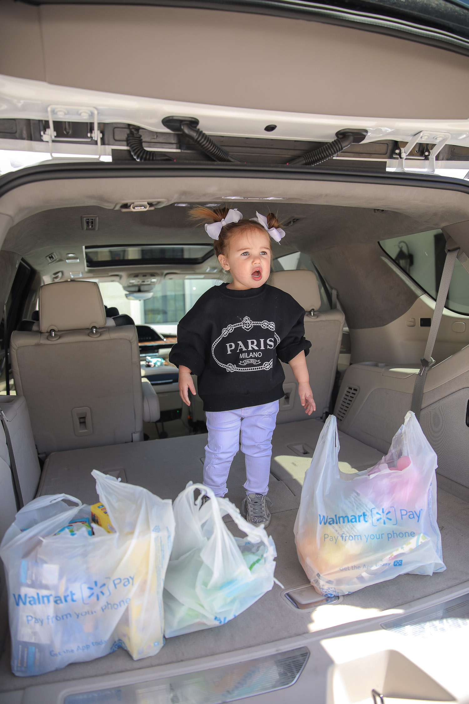 Easter Basket Ideas by popular US lifestyle blog, The Sweetest Thing: image of a little girl wearing a black Paris sweatshirt, white jeans, and white hair bows standing next to some Walmart shopping bags in the back of a White SUV.