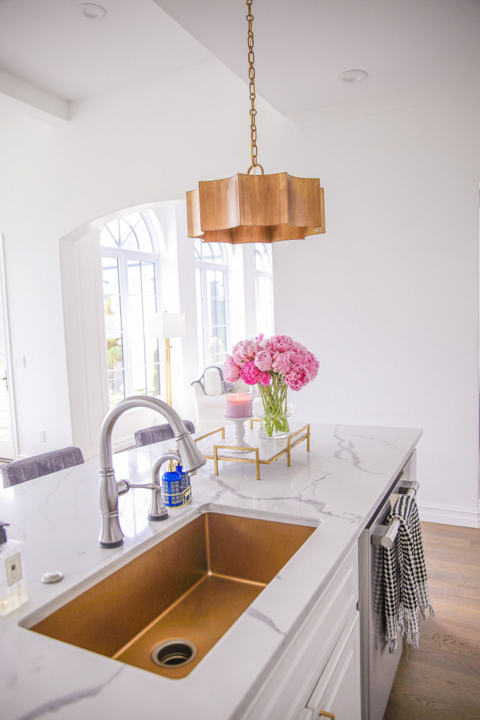 gold kitchen sink, touch2o faucet, Pinterest white kitchen gold hardware, white kitchen inspiration, Emily Gemma kitchen | Spring Kitchen by popular US life and style blog: image of a kitchen with white cabinets, wood flooring, wooden light pendants, marble counter tops, grey velvet and gold metal barstools, vase of pink peonies, and light pink and blue runner rug.