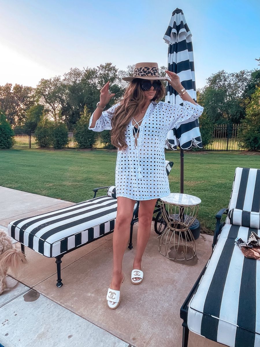 Summer outfit ideas, swimsuit, white lace cover up, flattering cover up, poolside style, summer 2021 |  Instagram Recap by popular US life and style blog, The Sweetest Thing: image of Emily Gemma wearing a straw boater hat with a leopard print scarf tied around it, white Chanel slide sandals, and a white eyelet swimsuit coverup with standing next to some poolside lounge chairs with black and white stripe cushions. 